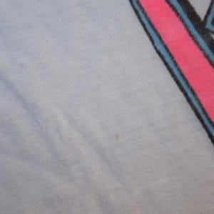 Op de afbeelding: Lichtblauwe stof met een roze en blauw grafisch ontwerp in de rechterbovenhoek. De afbeelding lijkt een gestileerde letter of symbool te zijn, met een zwarte omtrek en paarse accenten.