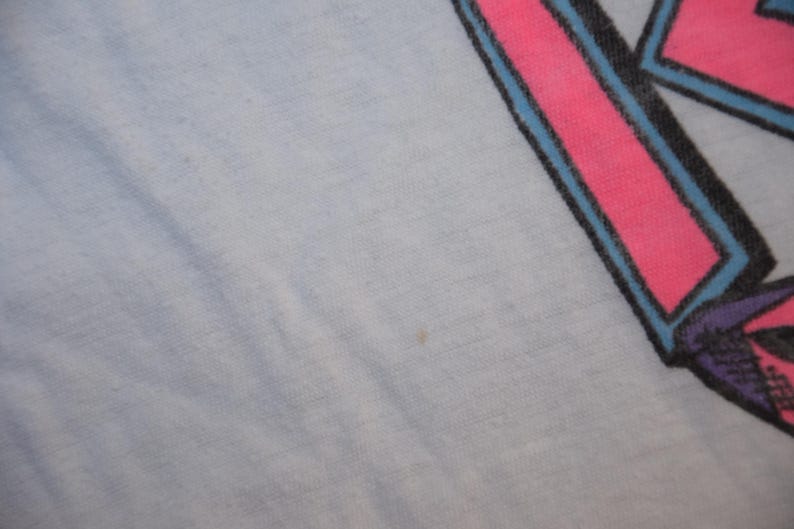 Op de afbeelding: Lichtblauwe stof met een roze en blauw grafisch ontwerp in de rechterbovenhoek. De afbeelding lijkt een gestileerde letter of symbool te zijn, met een zwarte omtrek en paarse accenten.