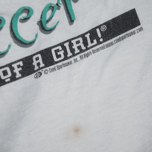 May include: White t-shirt with teal green text that reads "Soccer" and "Power of a Girl!" with a copyright symbol and the text "Coed Sportswear, Inc. All Rights Reserved www.coedsportswear.com"