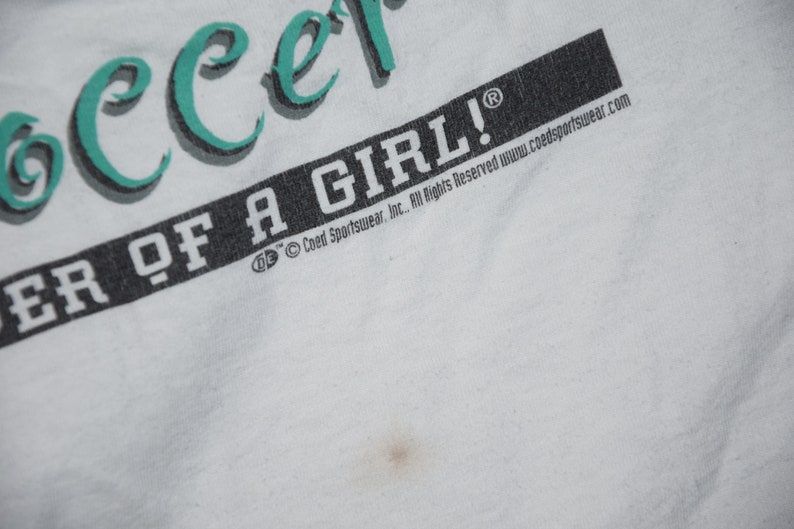 May include: White t-shirt with teal green text that reads "Soccer" and "Power of a Girl!" with a copyright symbol and the text "Coed Sportswear, Inc. All Rights Reserved www.coedsportswear.com"