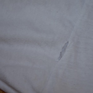Op de afbeelding: Close-up van witte stof met een scheur. De scheur onthult een grijs interieur. De stof lijkt van een t-shirt of een soortgelijk kledingstuk te komen. De textuur van de stof is zichtbaar.