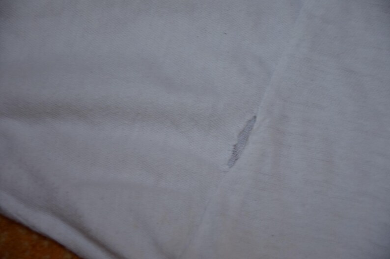 Op de afbeelding: Close-up van witte stof met een scheur. De scheur onthult een grijs interieur. De stof lijkt van een t-shirt of een soortgelijk kledingstuk te komen. De textuur van de stof is zichtbaar.