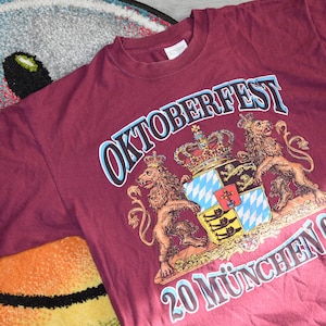 May include: A maroon t-shirt with a blue and white graphic of a coat of arms with two lions and the text "OKTOBERFEST 20 MÜNCHEN 02".