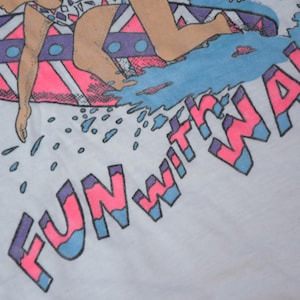 Op de afbeelding: Wit T-shirt met een retro ontwerp van een surfer op een golf. De tekst "FUN WITH WAVE" is roze en blauw. De surfer draagt een badpak. Het ontwerp heeft een vintage, jaren 80 stijl.