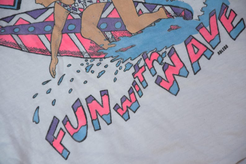 Op de afbeelding: Wit T-shirt met een retro ontwerp van een surfer op een golf. De tekst "FUN WITH WAVE" is roze en blauw. De surfer draagt een badpak. Het ontwerp heeft een vintage, jaren 80 stijl.