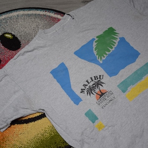 以下が含まれることがあります： カラフルなグラフィックが施されたヘザーグレーのTシャツ。「Malibu」の文字とヤシの木のイラスト、その下に「Caribbean White Rum Coconut」の文字が描かれています。デザインには青、緑、黄色の図形も含まれています。