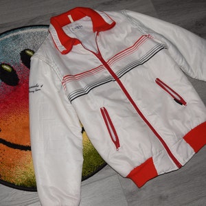 May include: White jacket with red and black stripes. The jacket has a zipper closure and two pockets. The text "remarkable" is printed on the left sleeve.