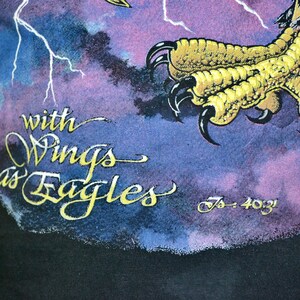 Może przedstawiać: Czarny t-shirt z graficznym wzorem przedstawiającym szpon orła i napis "with Wings as Eagles" złotymi literami. Tło ma fioletowo-niebieski wz&oacute;r chmur z błyskawicami.