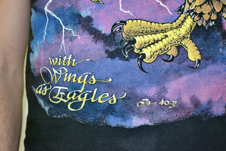 Może przedstawiać: Czarny t-shirt z graficznym wzorem przedstawiającym szpon orła i napis "with Wings as Eagles" złotymi literami. Tło ma fioletowo-niebieski wz&oacute;r chmur z błyskawicami.