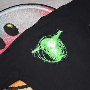以下が含まれることがあります： 黒いTシャツ。ネオングリーンのギターのグラフィックと、とげのある抽象的なデザインが特徴です。シャツは濃い色の生地でできており、模様のある表面に表示されています。