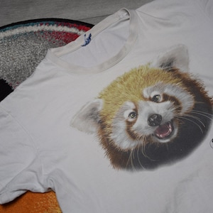May include: White t-shirt with a detailed illustration of a red panda's face. The red panda has brown and tan fur, with an open mouth. The shirt also has text and a logo that reads "Loro Parque Fundación We Care."