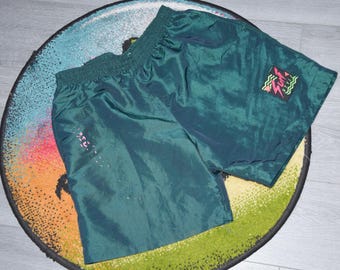 May include: Dark teal athletic shorts with an elastic waistband. The shorts have a small graphic on the right leg and a logo on the left leg. The shorts are made of a shiny, lightweight material.