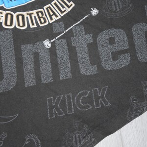 K&ouml;nnte beinhalten: Schwarzes T-Shirt mit einem wei&szlig;en und grauen Grafikdruck des Wortes "United" mit den W&ouml;rtern "Football" und "Kick" darunter. Die Grafik enth&auml;lt auch ein Wappen mit den W&ouml;rtern "Newcastle United" drumherum.