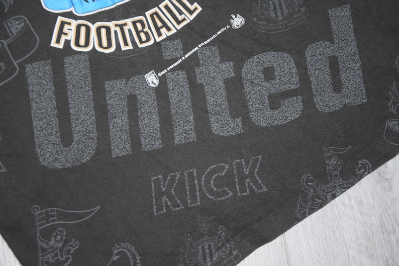 K&ouml;nnte beinhalten: Schwarzes T-Shirt mit einem wei&szlig;en und grauen Grafikdruck des Wortes "United" mit den W&ouml;rtern "Football" und "Kick" darunter. Die Grafik enth&auml;lt auch ein Wappen mit den W&ouml;rtern "Newcastle United" drumherum.
