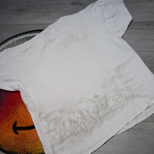 May include: White t-shirt with a gray graphic print of a group of people in a field. The graphic is on the back of the shirt.