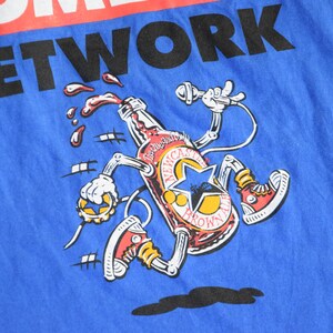 K&ouml;nnte beinhalten: Blaues T-Shirt mit einer Cartoon-Illustration einer Newcastle Brown Ale-Flasche. Die Flasche hat Arme, Beine und tr&auml;gt rote Turnschuhe. Der Text "COMEDY NETWORK" ist oben aufgedruckt.