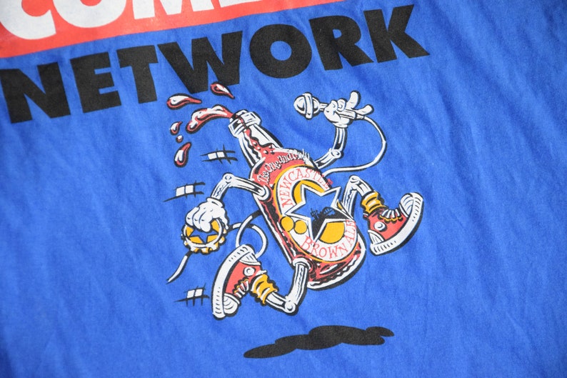 K&ouml;nnte beinhalten: Blaues T-Shirt mit einer Cartoon-Illustration einer Newcastle Brown Ale-Flasche. Die Flasche hat Arme, Beine und tr&auml;gt rote Turnschuhe. Der Text "COMEDY NETWORK" ist oben aufgedruckt.