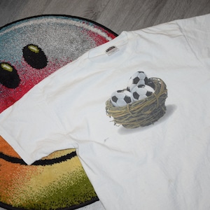 May include: White t-shirt with a graphic of soccer balls in a nest. The graphic is in shades of brown, black, and white.