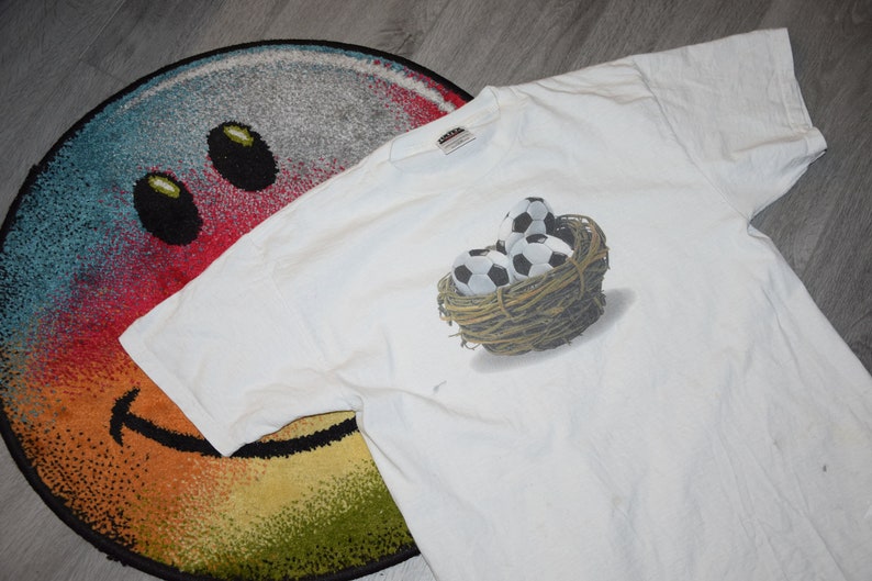 May include: White t-shirt with a graphic of soccer balls in a nest. The graphic is in shades of brown, black, and white.