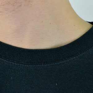 Op de afbeelding: Close-up van een zwart T-shirt met ronde hals. De kraag is geribbeld en de stof lijkt een zachte katoenmix te zijn. Het shirt is effen en heeft een eenvoudig, klassiek ontwerp.