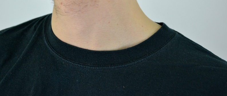 Op de afbeelding: Close-up van een zwart T-shirt met ronde hals. De kraag is geribbeld en de stof lijkt een zachte katoenmix te zijn. Het shirt is effen en heeft een eenvoudig, klassiek ontwerp.