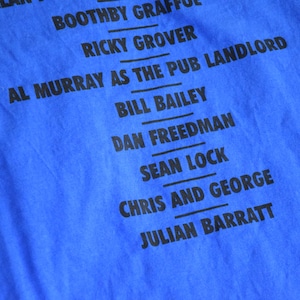 K&ouml;nnte beinhalten: Nahaufnahme eines leuchtend blauen T-Shirts mit schwarzem Text. Der Text listet Namen auf, darunter "AL MURRAY AS THE PUB LANDLORD" und "CHRIS AND GEORGE". Der Stoff hat eine sichtbare Textur.