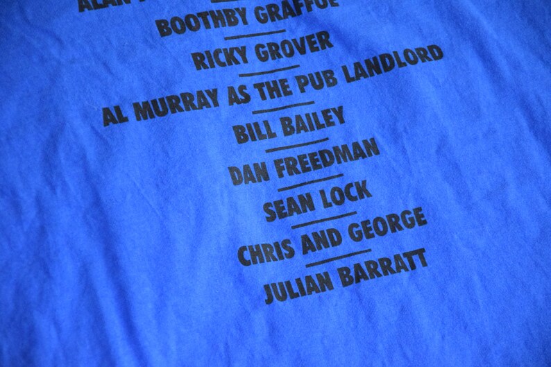 K&ouml;nnte beinhalten: Nahaufnahme eines leuchtend blauen T-Shirts mit schwarzem Text. Der Text listet Namen auf, darunter "AL MURRAY AS THE PUB LANDLORD" und "CHRIS AND GEORGE". Der Stoff hat eine sichtbare Textur.
