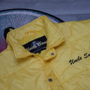 Könnte beinhalten: Hellgelbe Steppjacke mit Kragen und Druckknöpfen. Auf der Vorderseite der Jacke steht in schwarzer Schrift "Uncle Sam". Ein schwarzes Etikett mit Logo ist in der Nähe des Kragens zu sehen. Die Jacke hat die Größe M.