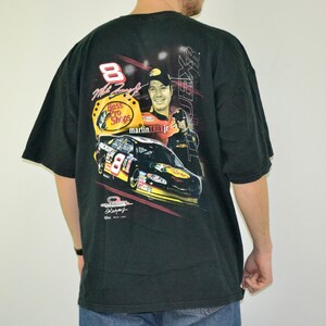 Op de afbeelding: Zwart t-shirt met een afbeelding van een raceauto, coureur en het Bass Pro Shops-logo. Het nummer 8 is zichtbaar. Het shirt heeft korte mouwen en een ronde hals.
