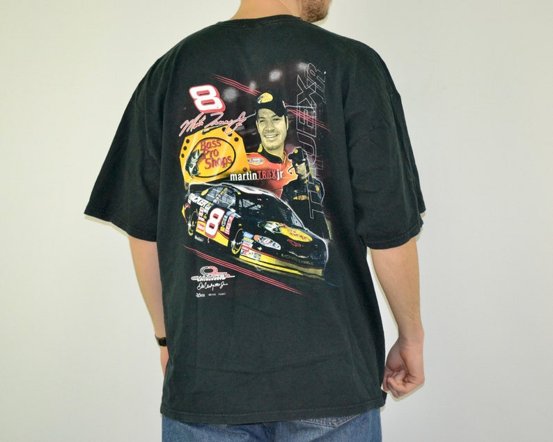 Op de afbeelding: Zwart t-shirt met een afbeelding van een raceauto, coureur en het Bass Pro Shops-logo. Het nummer 8 is zichtbaar. Het shirt heeft korte mouwen en een ronde hals.