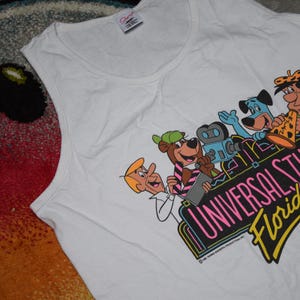 Może przedstawiać: Biały podkoszulek z kolorową grafiką postaci z kreskówek i napisem "UNIVERSAL STUDIOS Florida". Projekt zawiera postacie takie jak Fred Flinstone i Yogi Bear. Koszulka w stylu vintage.