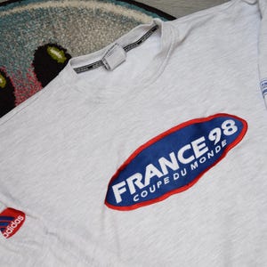 Puede incluir: Camiseta de manga larga gris claro con un parche ovalado azul y rojo grande que dice "FRANCE 98 COUPE DU MONDE". La camiseta también presenta el logotipo de Adidas y un pequeño parche con el mismo texto. Vintage.