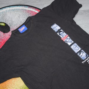 Puede incluir: Camiseta negra con un gráfico de seis retratos y el texto "Thunderbirds are GO!". La camiseta tiene cuello redondo y mangas cortas. Una etiqueta azul es visible cerca del cuello.