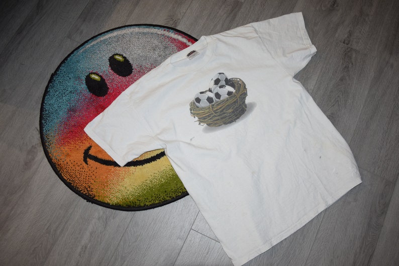 May include: White t-shirt with a graphic of a nest of soccer balls. The nest is made of twigs and the soccer balls are black and white.