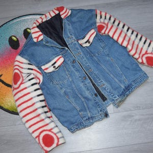 May include: A blue denim jacket with red and white striped sleeves and accents. The jacket has a black lining and button closures. A colorful smiley face rug is in the background.
