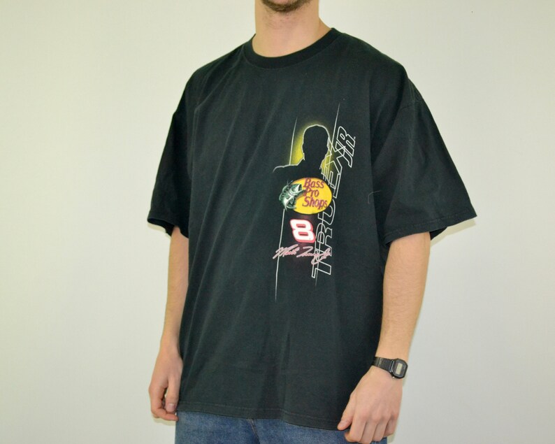 Op de afbeelding: Zwart T-shirt met korte mouwen met een grafisch ontwerp met de tekst "TRUE AIR" en "Bass Pro Shops". Het ontwerp bevat een silhouet en het nummer 8. Het shirt is gemaakt van zacht materiaal.