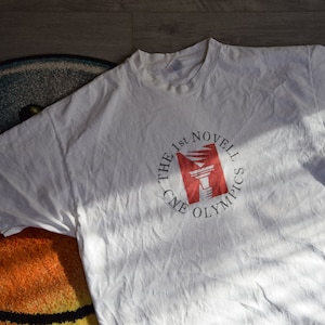 Op de afbeelding: Wit T-shirt met een rode en witte cirkelvormige afbeelding met de tekst "The 1st Novel CNE Olympics".