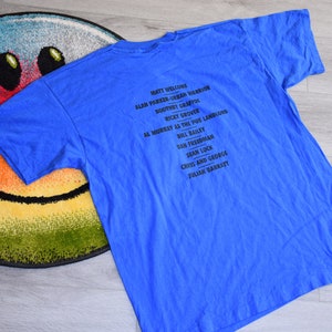 K&ouml;nnte beinhalten: Ein blaues T-Shirt mit einer schwarzen Namensliste. Das Shirt liegt auf einem runden Teppich mit einem Smiley-Gesicht. Der Text enth&auml;lt Namen wie Matt Welcome und Alan Parker-Urban Warrior.