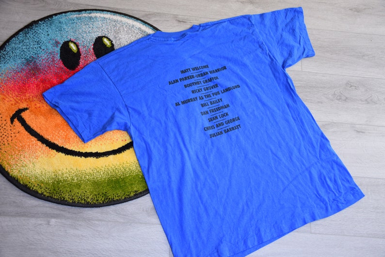 K&ouml;nnte beinhalten: Ein blaues T-Shirt mit einer schwarzen Namensliste. Das Shirt liegt auf einem runden Teppich mit einem Smiley-Gesicht. Der Text enth&auml;lt Namen wie Matt Welcome und Alan Parker-Urban Warrior.