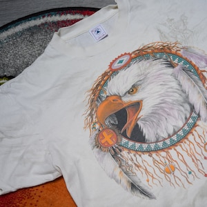 May include: White t-shirt with a graphic of a bald eagle with a Native American dreamcatcher design. The eagle has a white head and body with orange and brown feathers. The dreamcatcher is made of beads and feathers in shades of blue, orange, and brown.