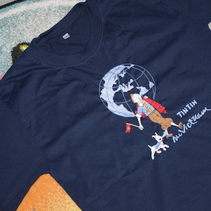 May include: Navy blue t-shirt featuring an embroidered design of Tintin, a globe, and Snowy the dog. The design includes the text "Tintin au Vietnam". The shirt has a crew neck and short sleeves.