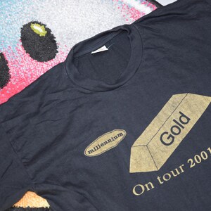 May include: Black t-shirt with a gold bar graphic and the text "Gold" and "On tour 2001". The t-shirt also has a small oval with the text "millennium" on it.