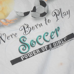 May include: White t-shirt with a graphic of a yellow chick kicking a soccer ball. The text reads "We're Born to Play Soccer POWER OF A GIRL!"