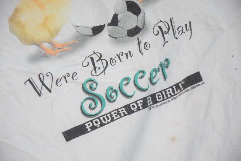 May include: White t-shirt with a graphic of a yellow chick kicking a soccer ball. The text reads "We're Born to Play Soccer POWER OF A GIRL!"