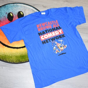 K&ouml;nnte beinhalten: Ein blaues T-Shirt mit dem Text "Newcastle Brown Ale National Comedy Network" in Rot, Wei&szlig; und Schwarz. Unter dem Text ist eine Cartoonfigur abgebildet. Das Shirt liegt auf einem bunten Smiley-Teppich.