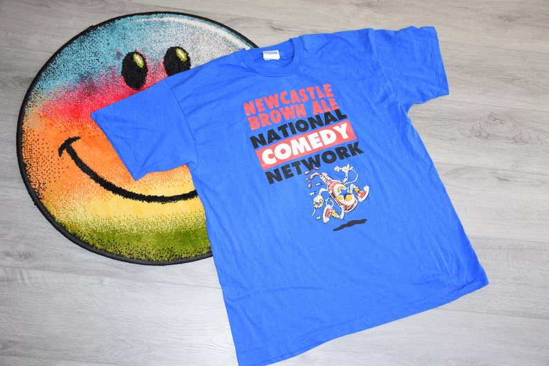 K&ouml;nnte beinhalten: Ein blaues T-Shirt mit dem Text "Newcastle Brown Ale National Comedy Network" in Rot, Wei&szlig; und Schwarz. Unter dem Text ist eine Cartoonfigur abgebildet. Das Shirt liegt auf einem bunten Smiley-Teppich.