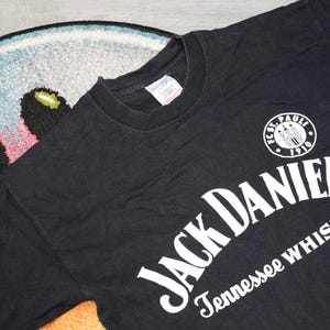Puede incluir: Camiseta negra con el logotipo de Jack Daniel's Tennessee Whiskey en blanco. También es visible un logotipo circular blanco con texto. La camiseta está hecha de un material suave.