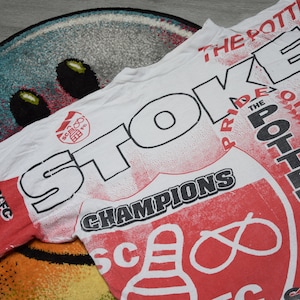 May include: White t-shirt with a red and white all-over print. The shirt features the words "Stoke", "Champions", "Pride of the Potteries", and "SCFC".