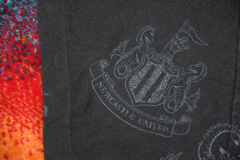 K&ouml;nnte beinhalten: Schwarz-wei&szlig;es Grafikdruck des Wappens des Fu&szlig;ballvereins Newcastle United. Das Wappen zeigt einen Schild mit drei vertikalen Streifen, zwei Seepferdchen und ein Banner mit der Aufschrift "Newcastle United".