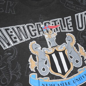 K&ouml;nnte beinhalten: Schwarzes T-Shirt mit einem wei&szlig;-schwarzen Logo des Newcastle United Football Clubs. Das Logo zeigt ein Wappen mit zwei Seepferden, einem Schloss und einem Banner mit der Aufschrift "Newcastle United".
